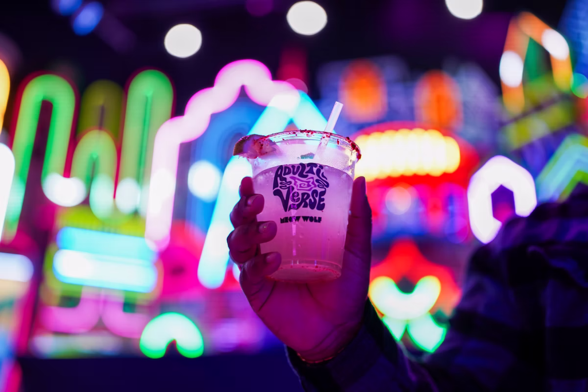 A hand holding an adultiverse cup with a drink in front of neon lights at Meow Wolf Grapevine