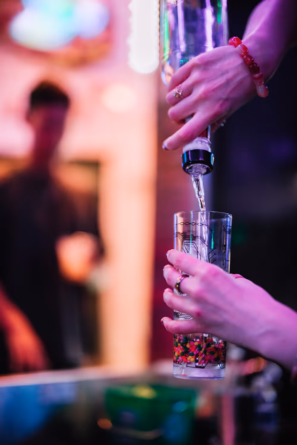 Hands pouring a clear liquid into a glass filled with colorful candy or beads in a dim, colorful setting.