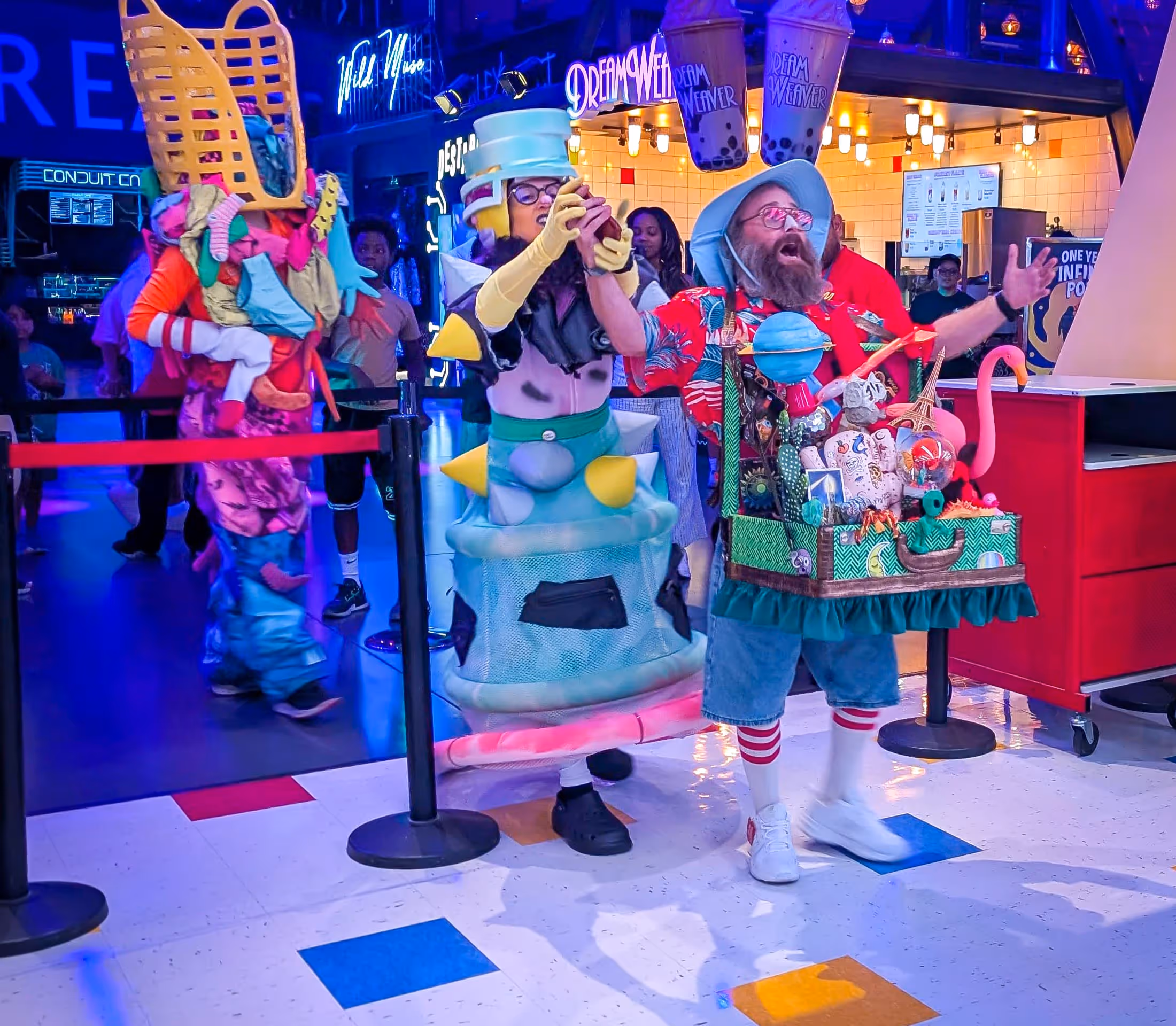 Three people in colorful, imaginative costumes posing indoors at a lively event with neon signs.